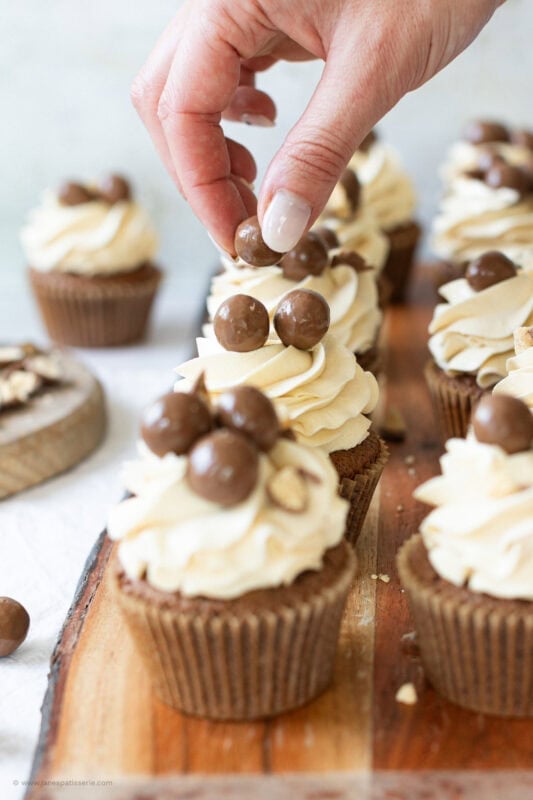 A hand placing maltesers on a decorated cupcake