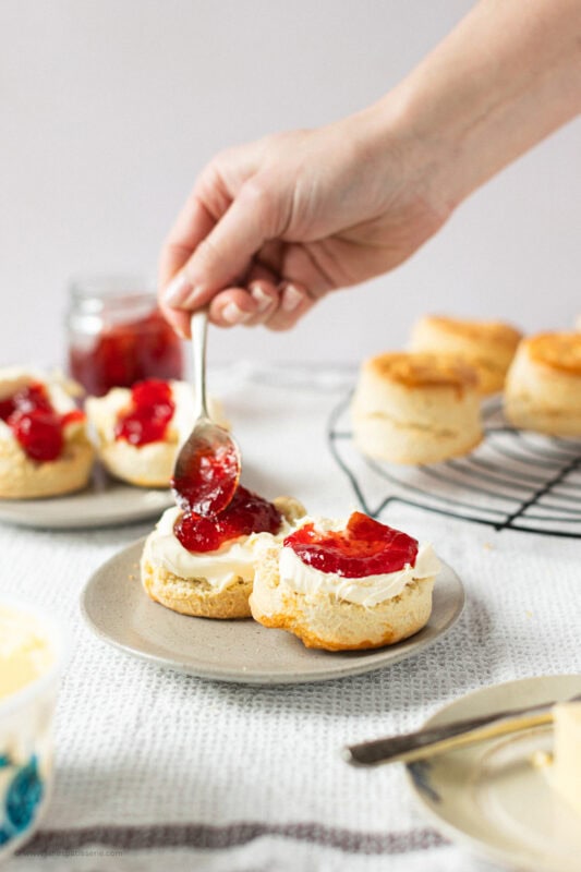 A hand spooning jam onto a buttered classic buttery scone