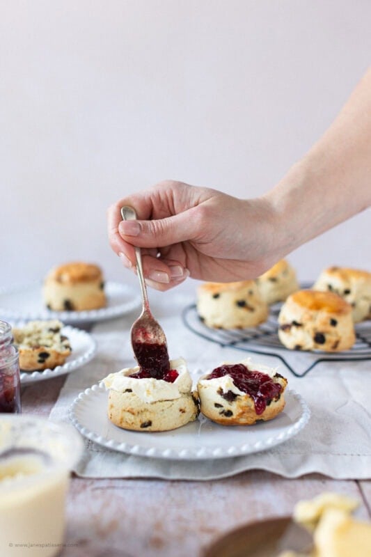 A spoon decorating a cut Fruit Scone with Clotted Cream and jam